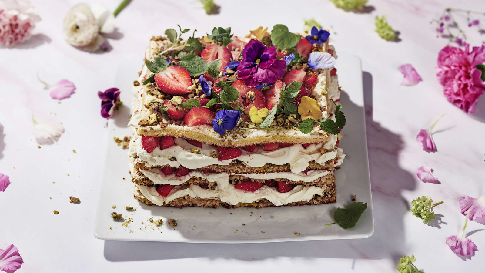 Pistaasi-britakakku, joka on koristelut orvokeilla, mansikoilla ja mintunlehdillä. Kakun ympärillä vaaleanpunaisia kukkia ja kukkien terälehtiä.