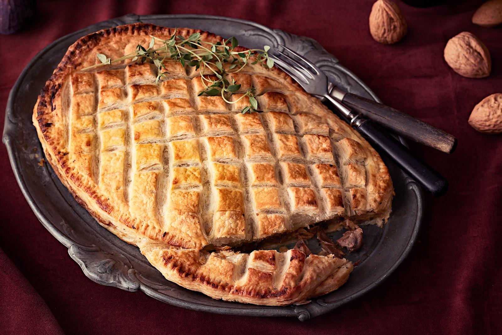 Vegaaninen Wellington metallisella tarjoiluvadilla. Vadin reunalla tarjoiluottimet ja vieressä kuorellisia saksanpähkinöitä.