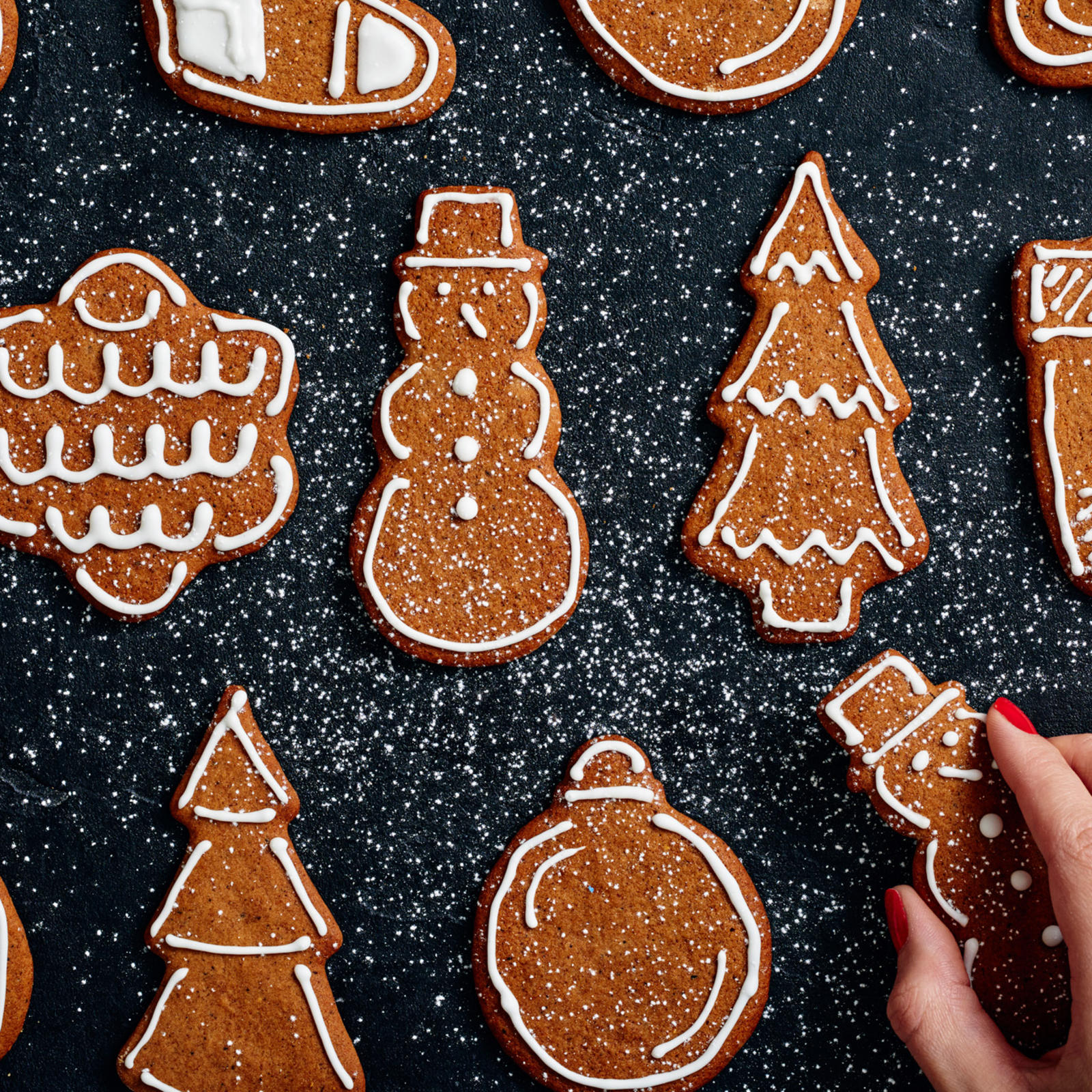 Koristeltuja pipareita mustalla alustalla. Alareunassa käsi nappaamassa piparin.