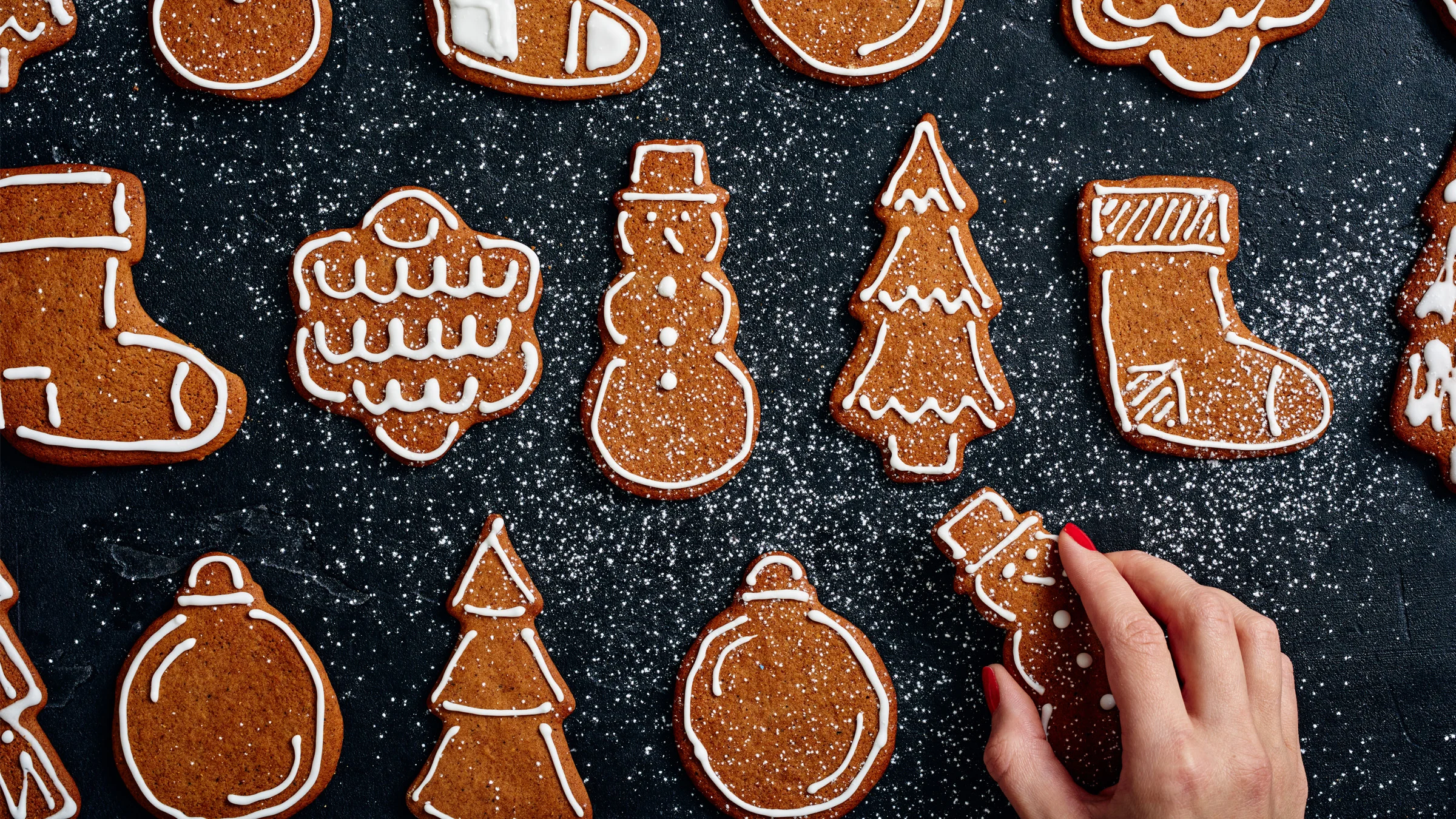 Koristeltuja pipareita mustalla alustalla. Alareunassa käsi nappaamassa piparin.