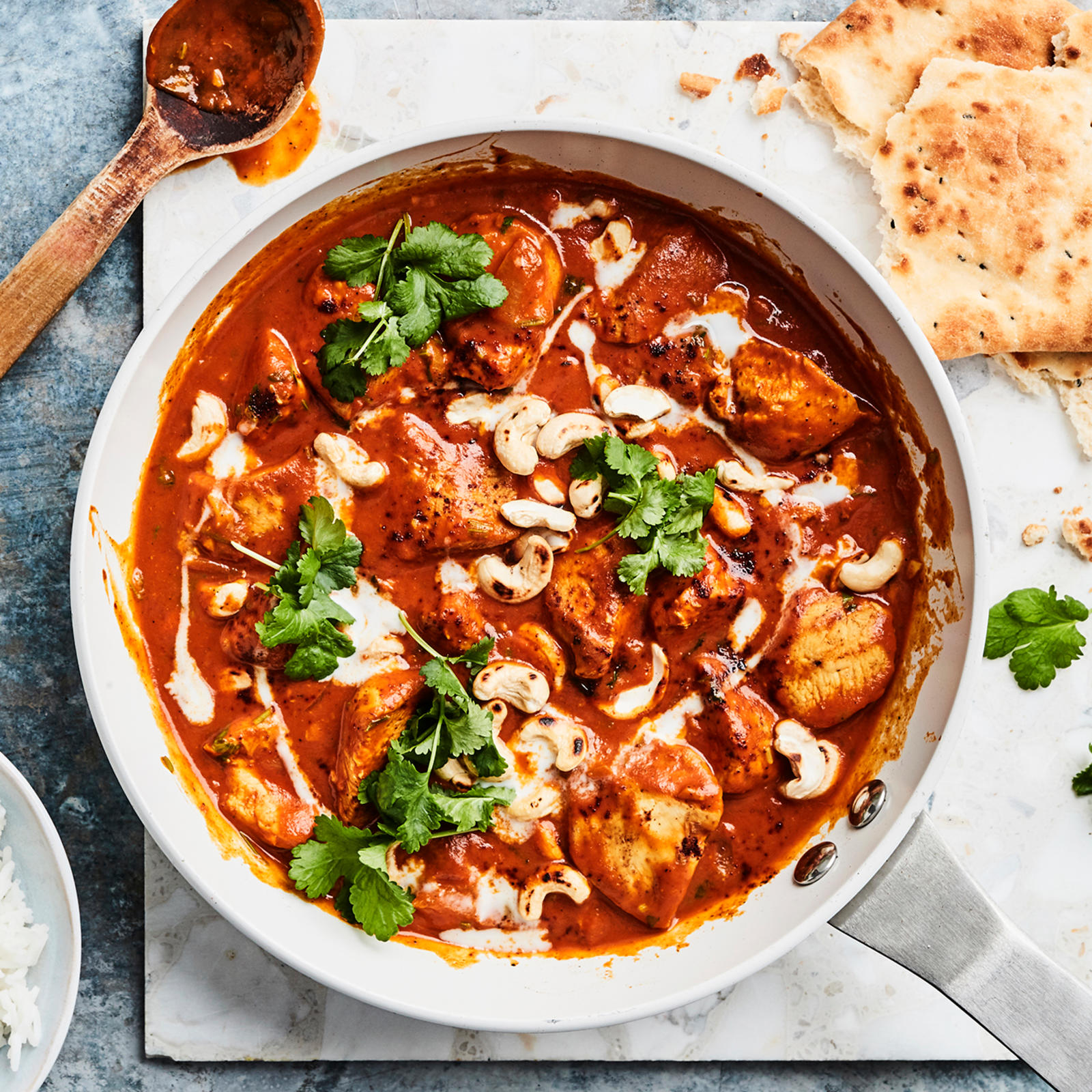 Butter chicken teräksisessä paistinpannussa. Vieressä puinen kauha, murrettua naanleipää, korianteria ja kulhollinen riisiä.