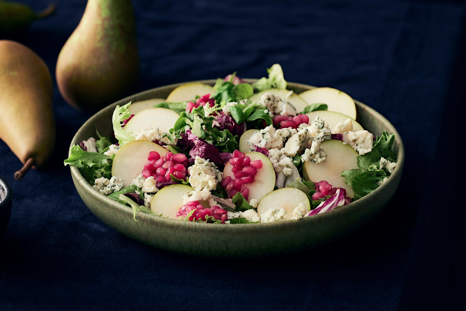Päärynä-sinihomejuustosalaattia vihreässä kulhossa. Salaatin päällä granaattiomenakastiketta ja salaatin vieressä päärynöitä sekä granaattiomenakastiketta kulhossa.