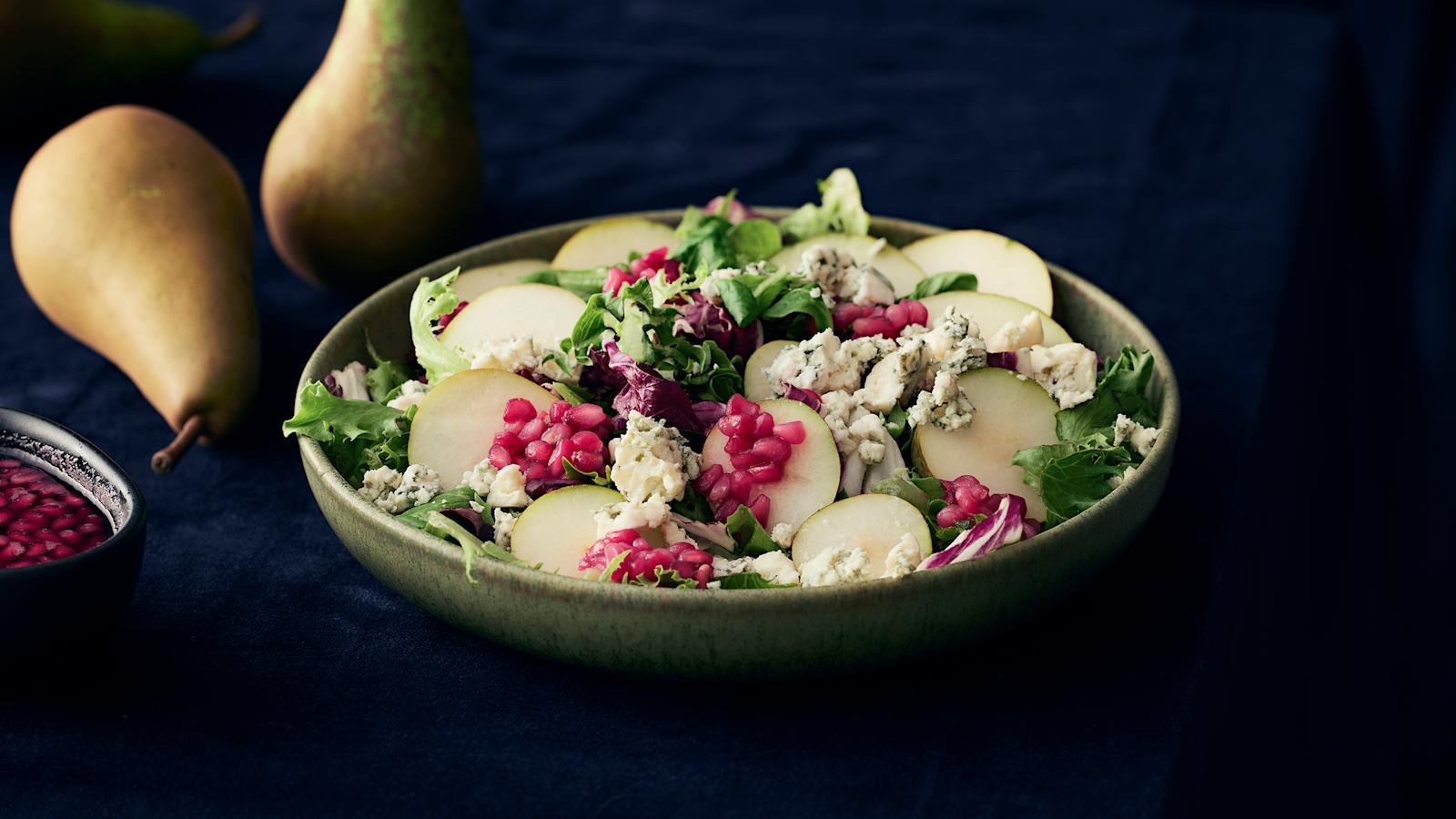 Päärynä-sinihomejuustosalaattia vihreässä kulhossa. Salaatin päällä granaattiomenakastiketta ja salaatin vieressä päärynöitä sekä granaattiomenakastiketta kulhossa.