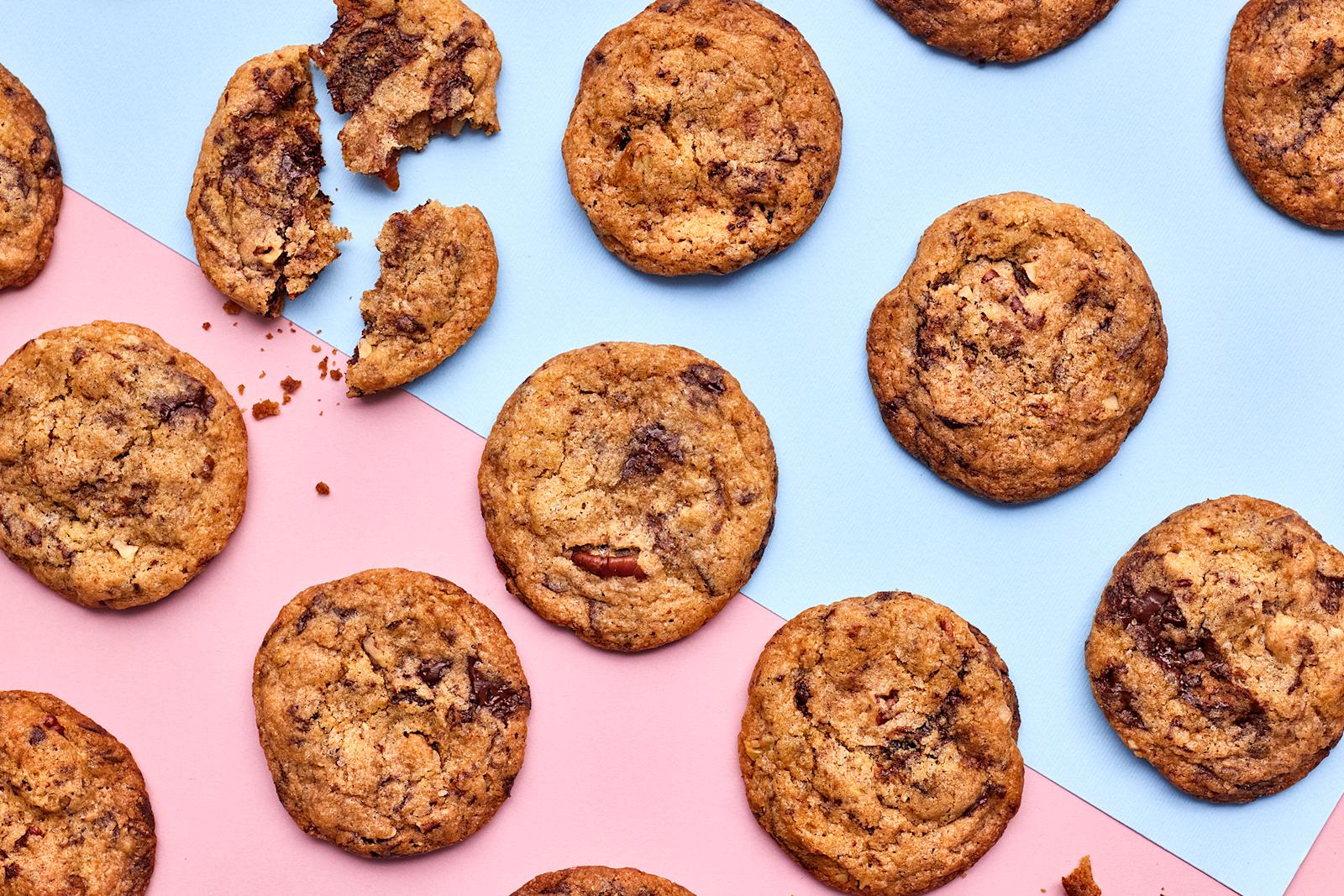 Vegaanisia suklaacookieita vaaleanpunaisella ja vaaleansinisellä taustalla. Yksi cookie murrettu kolmeen osaan.