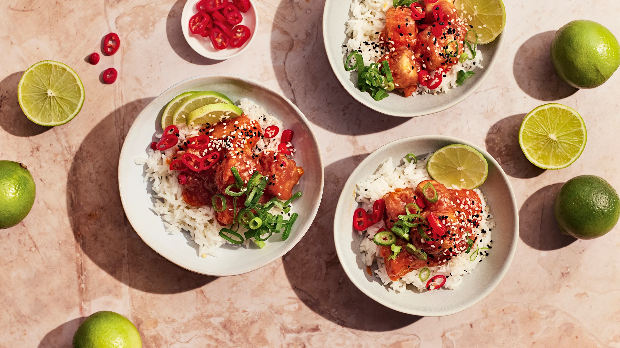 Sticky tofu ja riisiä kolmessa kulhossa limellä, chilillä, kevätsipulilla ja seesaminsiemenillä koristeltuna. Vieressä limejä ja chiliviipaleita.