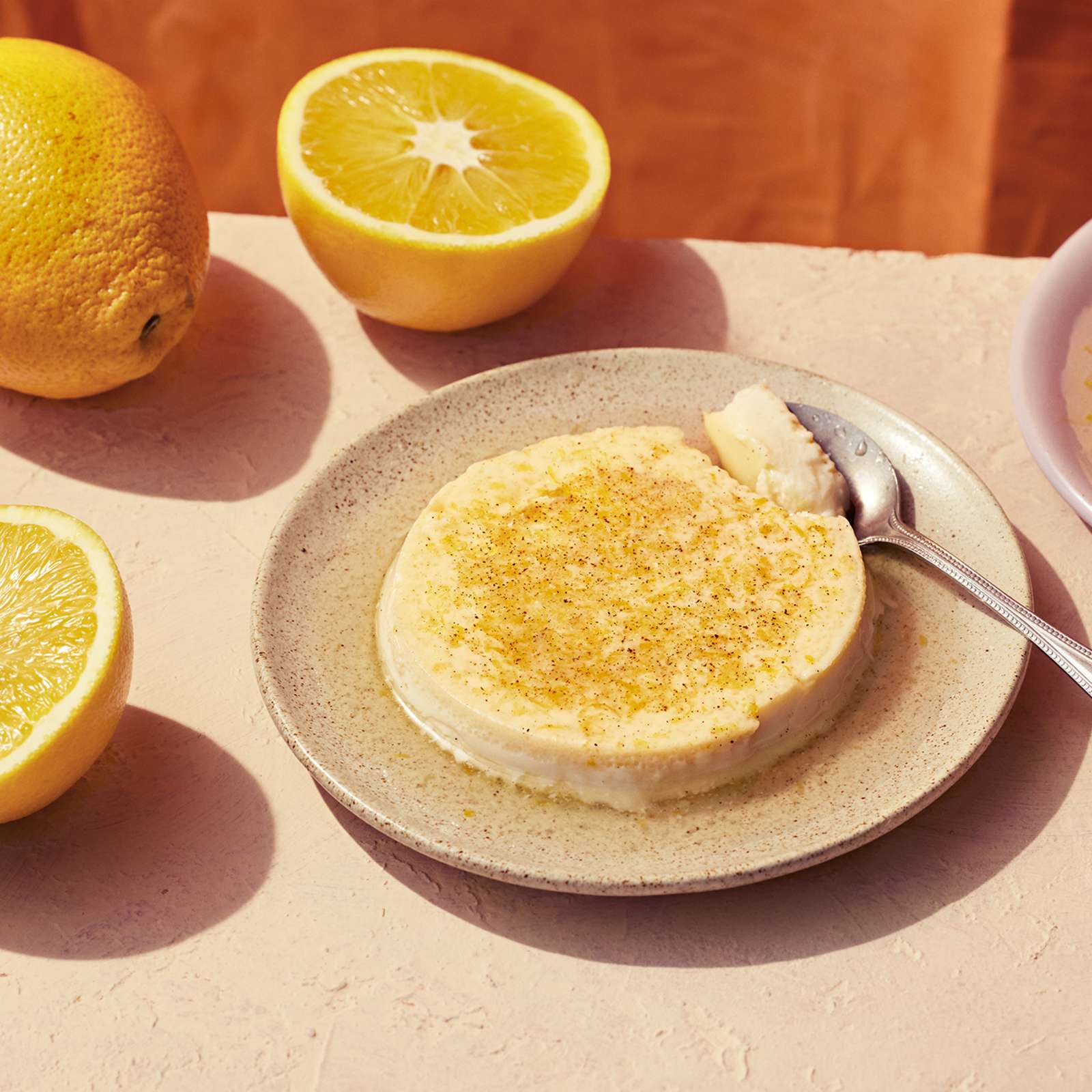 Appelsiininen flan eli paahtovanukas lautasella, jossa lusikka. Vieressä toinen vanukas ja puolitettuja appelsiineja. 