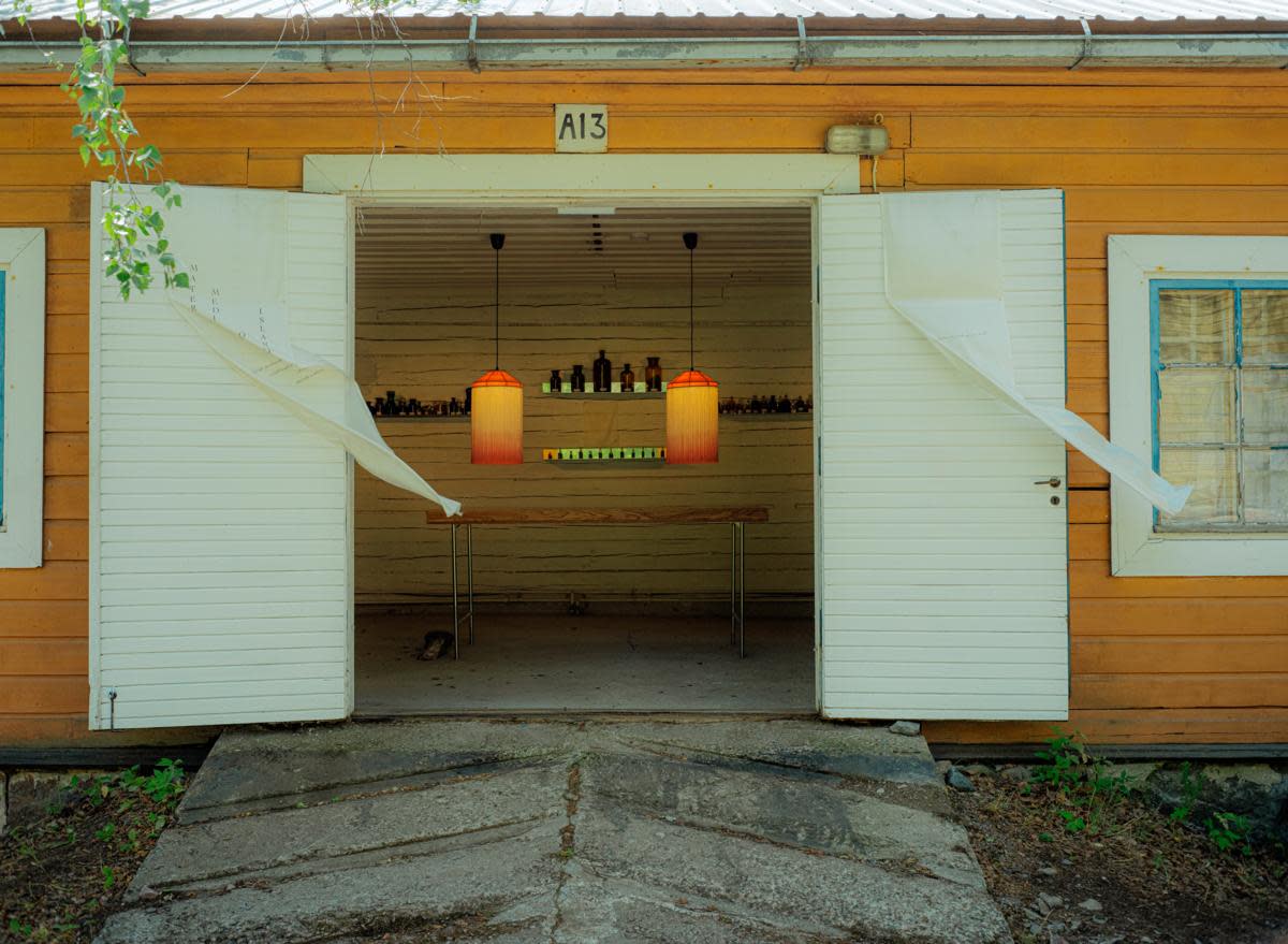 Keltaisen puutalon valkoiset pariovet ovat auki. Sisällä näkyy pöytä ja kaksi oranssia lamppua.