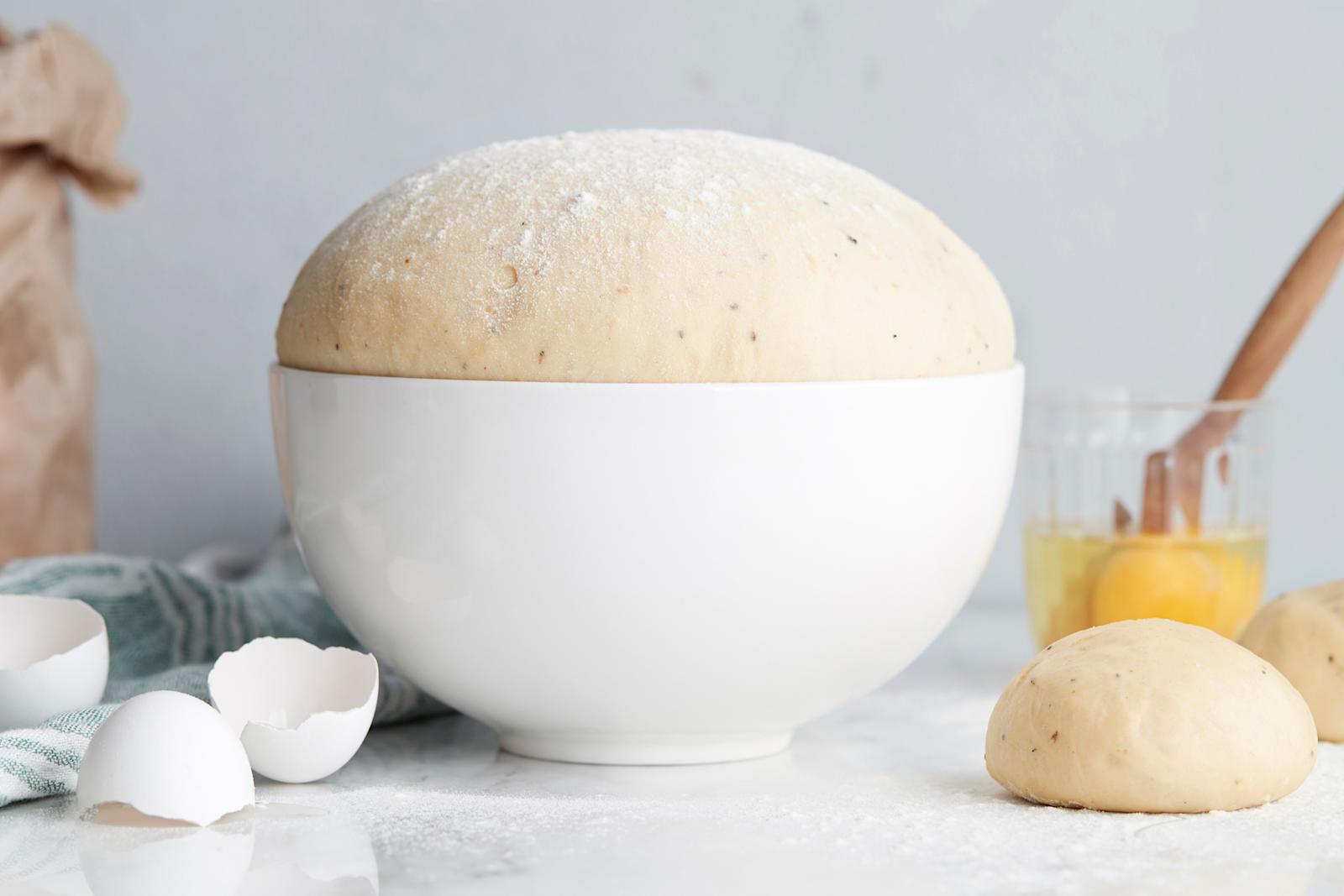 Kohonnut pullataikina valkoisessa taikinakulhossa, kaksi taikinasta leivottua pikkupullaa, lasiin rikottu kananmuna ja kananmunan kuoria vaalealla pöydällä.