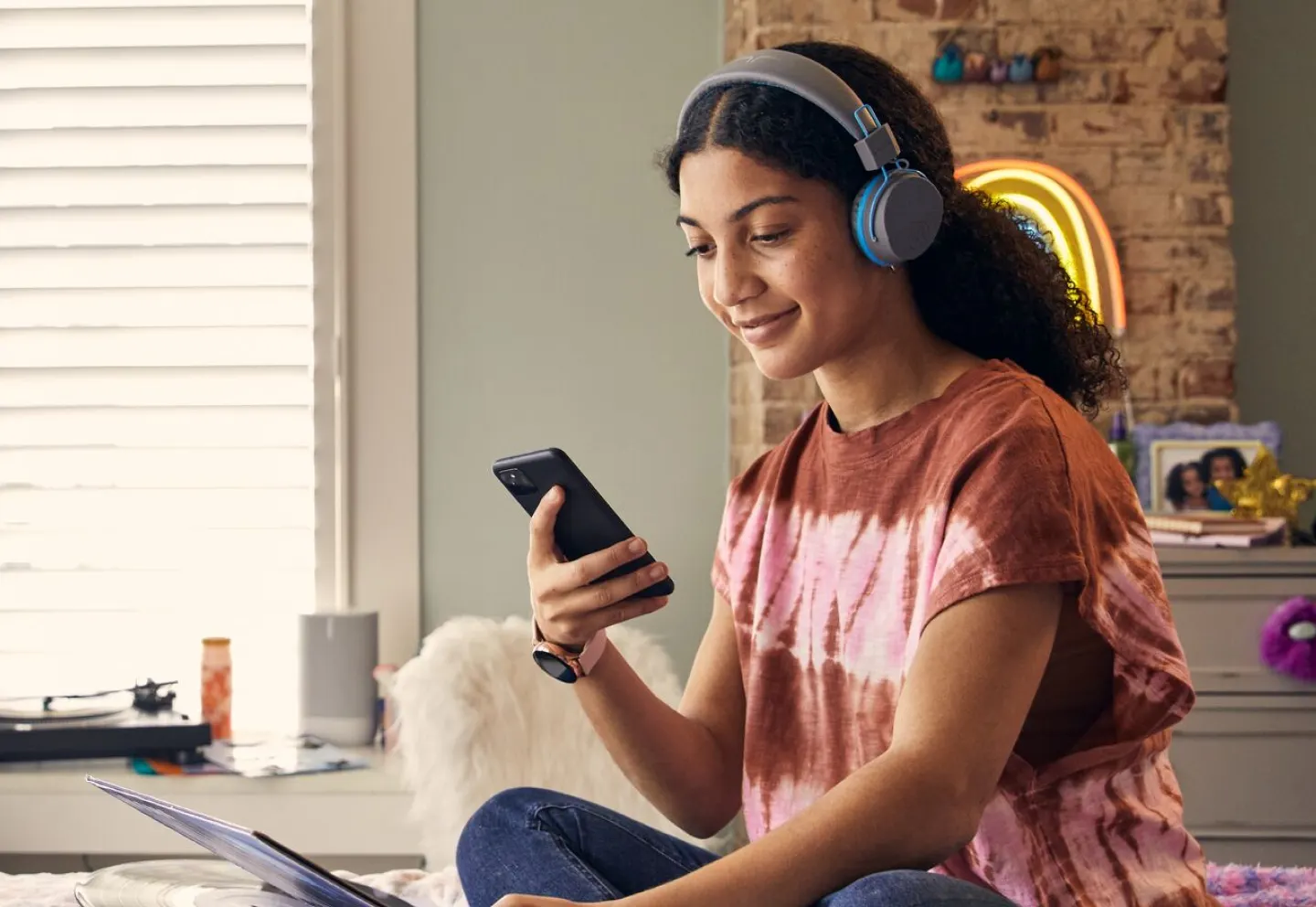 Girl with headphones on sitting a bed using a phone and laptop
