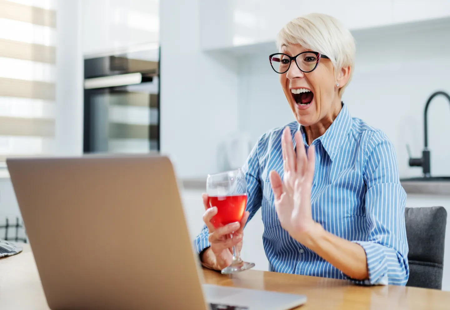 An older woman holding a drink in one hand and using a laptop