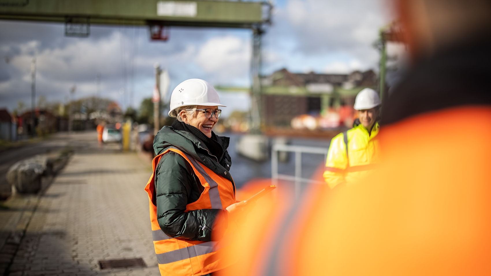 En kvinne med hjelm og refleksvest smiler mens hun leder et prosjekt på en byggeplass.
