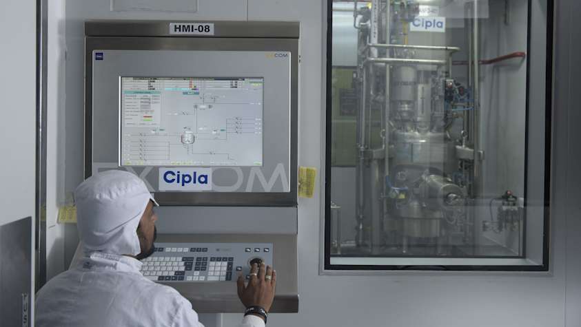 A person is standing in front of a white wall, wearing a black shirt and holding a black object in their hand.