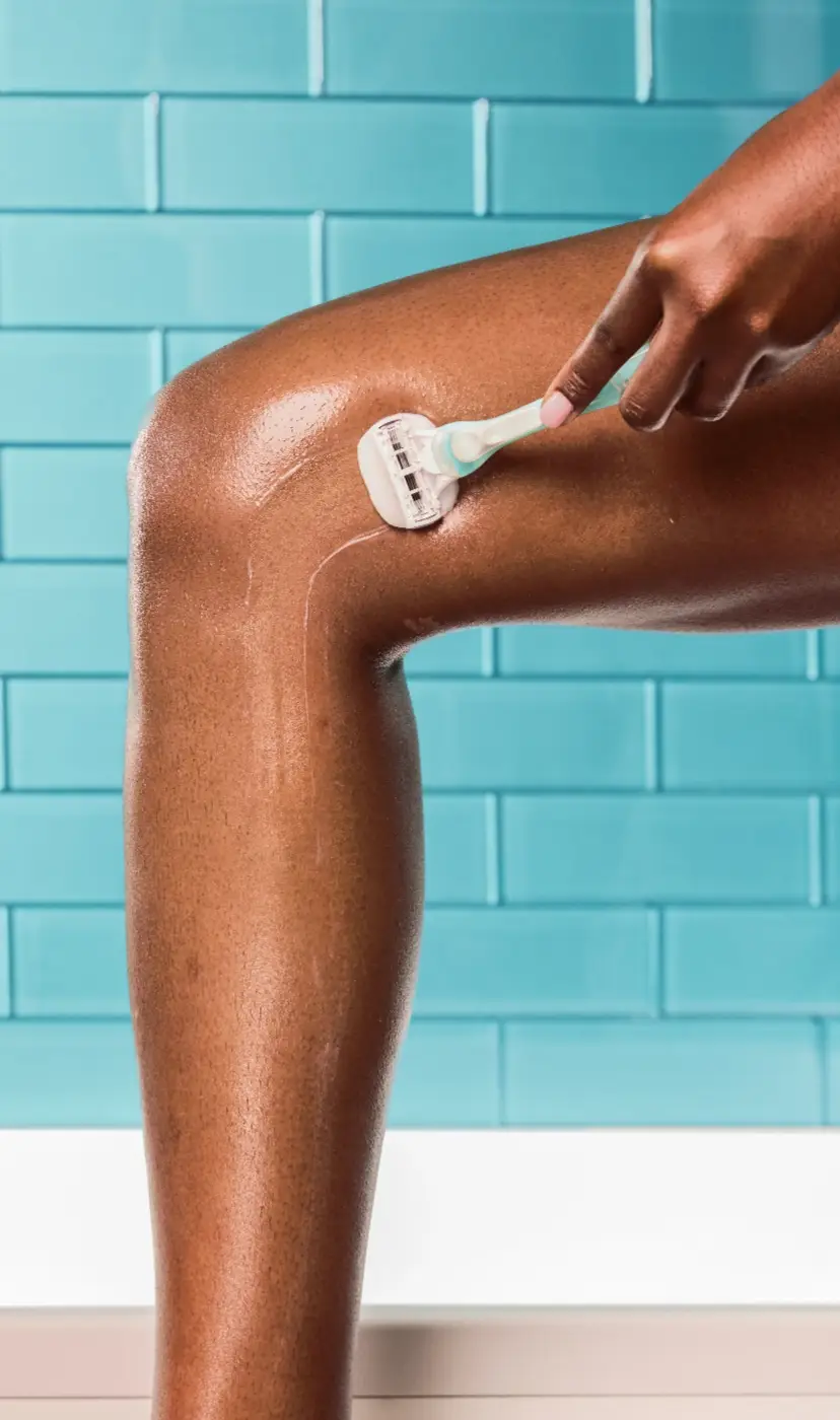 Woman in a blue bathroom shaving her left thigh with a ComfortGlide razor