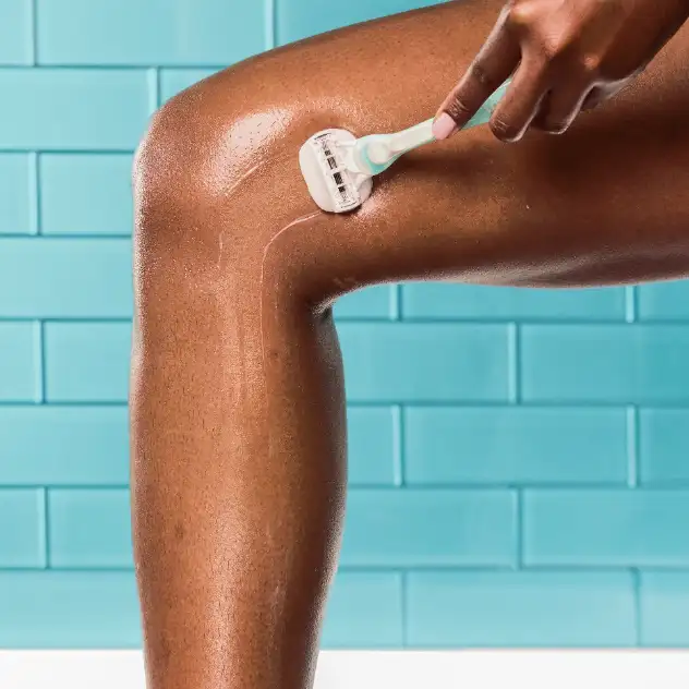 Woman using a razor to shave a lower leg against a tiled background