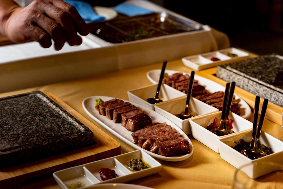 A hand reaches over a table with a variety of grilled meat slices arranged on white trays. Each tray has sauce-filled compartments with small spoons. A hot stone sits nearby, ready to cook more meat. A tablecloth and napkin are visible.