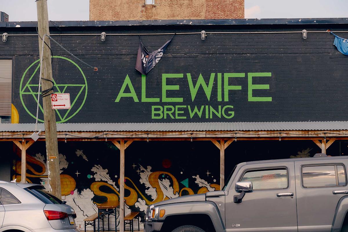 The exterior of Alewife Brewing, with a large green sign on a dark brick wall and murals below. A Jeep and other parked cars are in front, and a "No Parking" sign is attached to a pole.