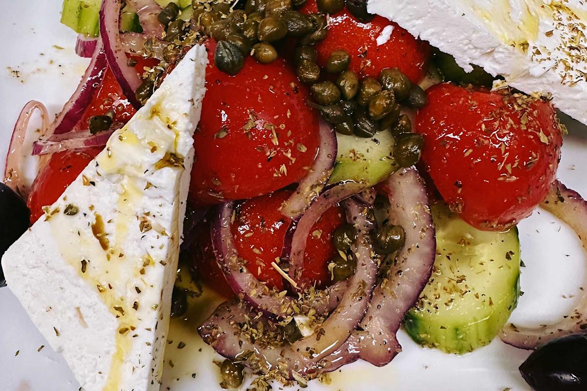 A close-up of a Greek salad with tomato, cucumber, red onion, black olives, capers, feta cheese slices, and oregano, drizzled with olive oil on a white plate.