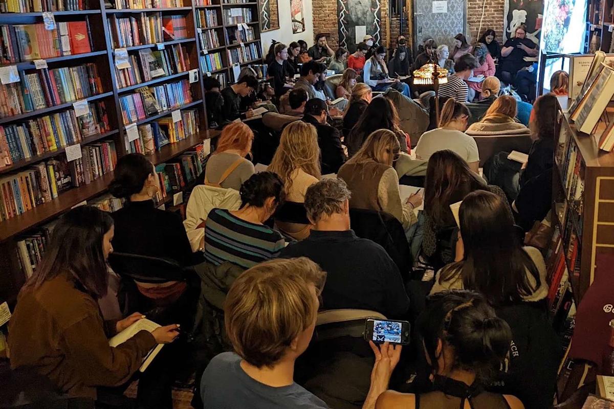 A crowded room in a bookstore with people seated and standing, attentively listening to a speaker. Bookshelves line the walls, and some attendees are taking notes or looking at their phones. The setting is cozy and engaging.