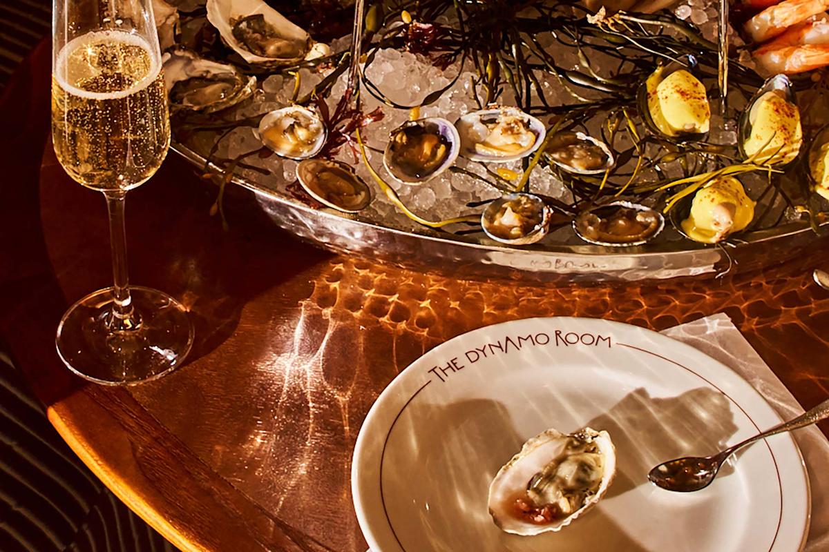 A glass of champagne and a plate with a single oyster sit on a wooden table beside a large ice-filled seafood platter with oysters, clams, and lemon wedges at The Dynamo Room restaurant.