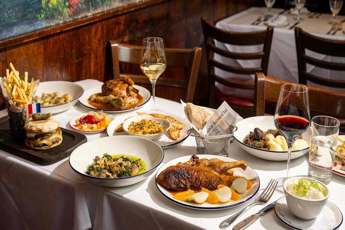 A table set for two with plates of French cuisine, including roast chicken, burgers, fries, vegetables, bread, wine glasses with red and white wine, and side dishes in a cozy restaurant setting.