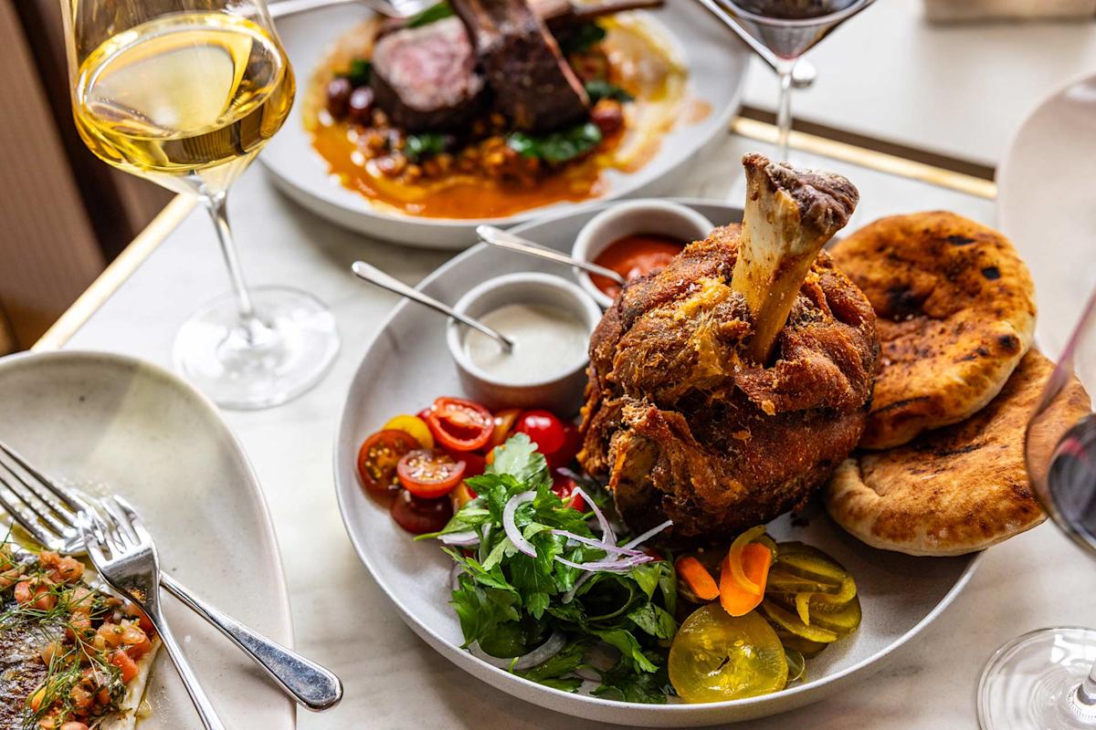 A plate with crispy roast meat on the bone, fresh herbs, sliced cherry tomatoes, pickled vegetables, sauces, and flatbread, surrounded by glasses of wine and additional dishes on a table.