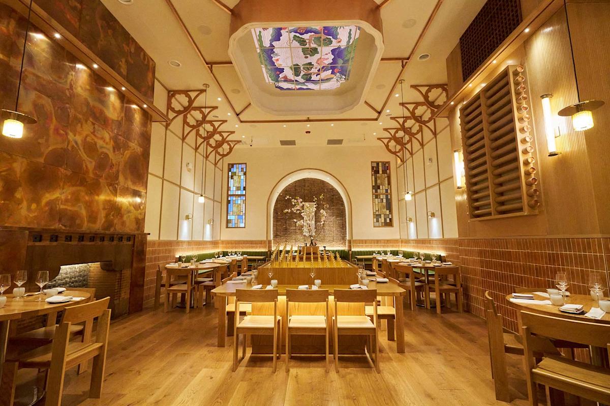 A modern restaurant interior with wooden tables and chairs, warm lighting, and an artistic ceiling mural. The room features a central arched wall with a textured art display and a spacious, inviting atmosphere.