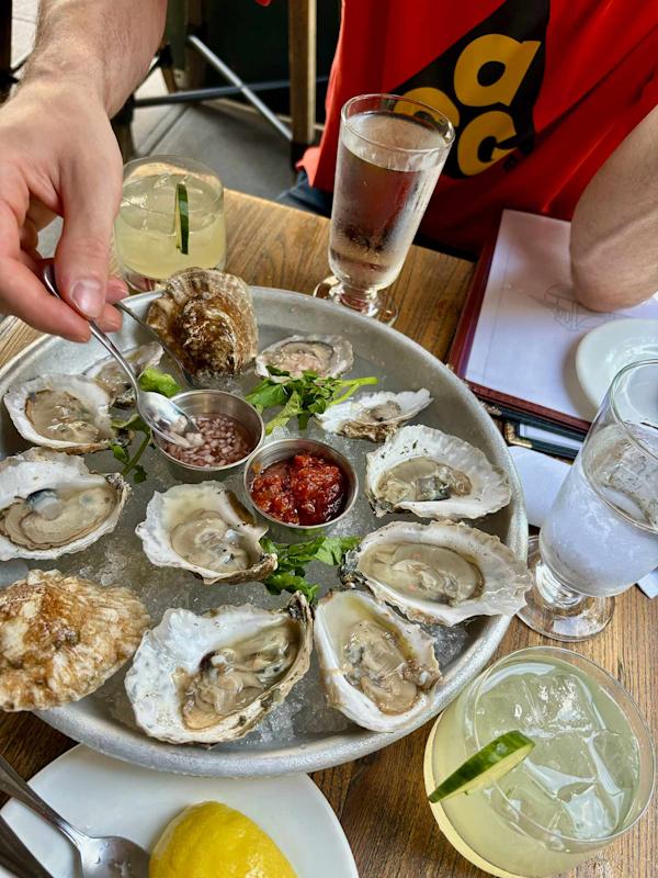 A person spoons sauce onto fresh oysters arranged on a round platter with lemon and garnishes, surrounded by drinks and a partially visible person in a red shirt at a restaurant table.