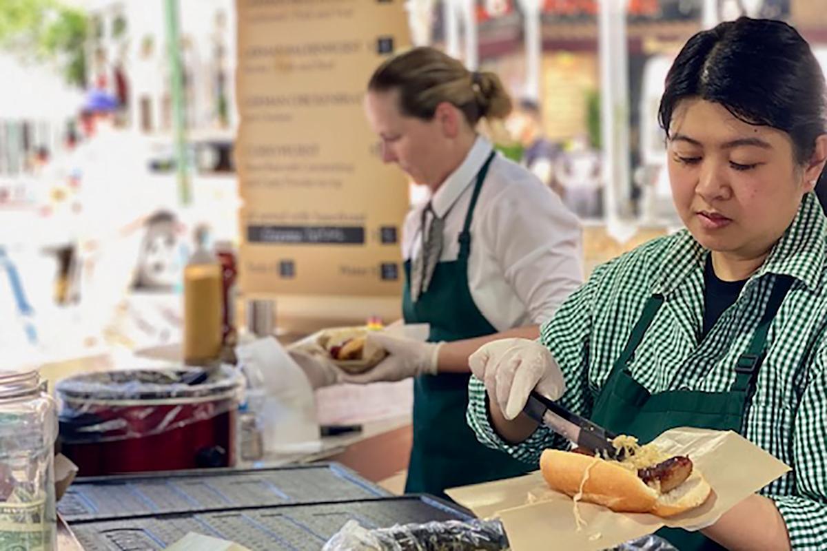 Two women in green aprons prepare food at an outdoor stand. One places sausage in a bun with tongs, while the other prepares another dish in the background. Condiments and a menu board are visible on the counter.