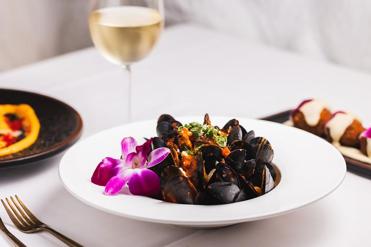 A white plate of mussels garnished with herbs and a purple orchid flower, set on a white tablecloth with a glass of white wine and other dishes in the background.
