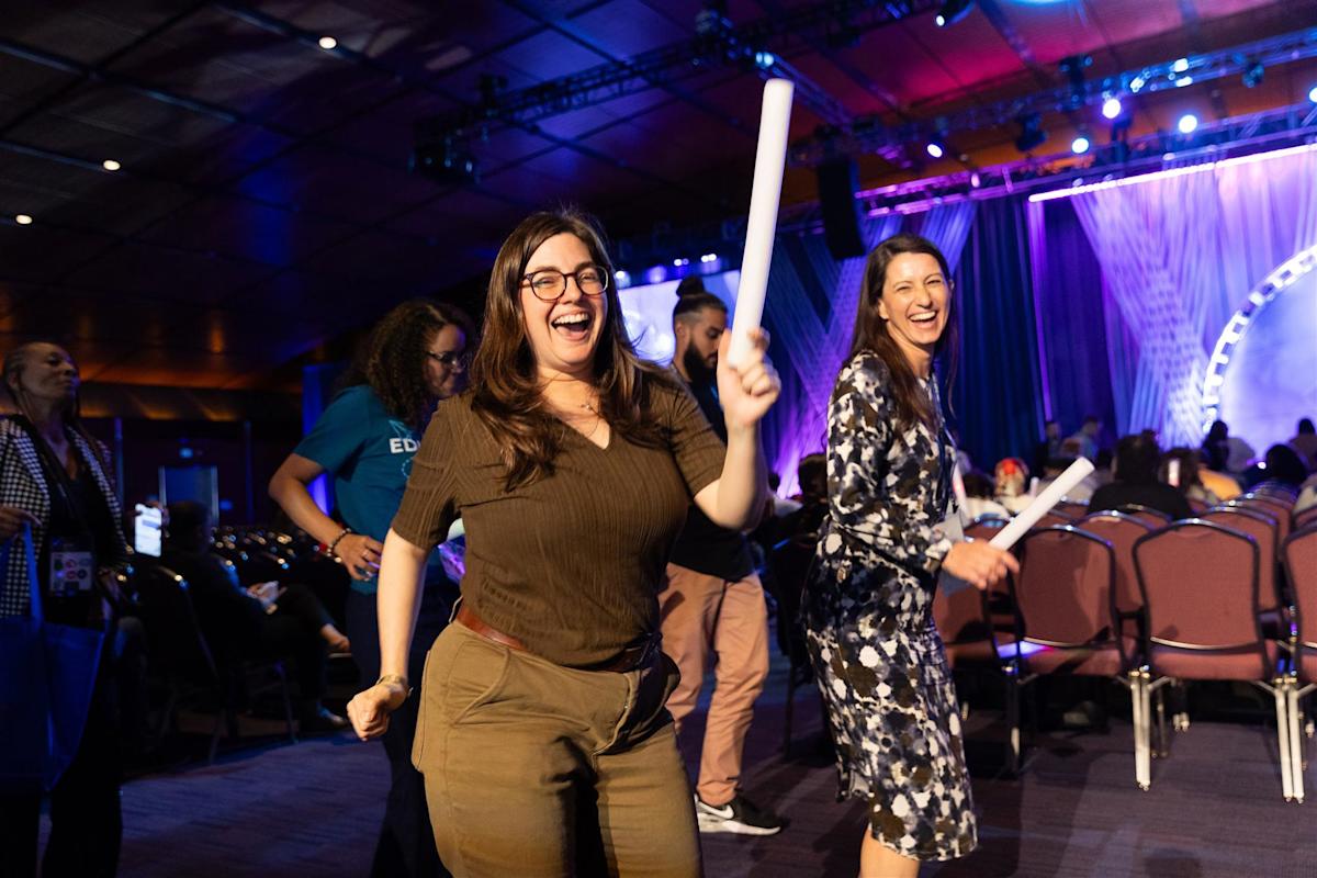 Two women are joyfully dancing with light sticks at an indoor event. The room is filled with people seated and standing. Bright stage lights and purple curtains create a festive atmosphere in the background.
