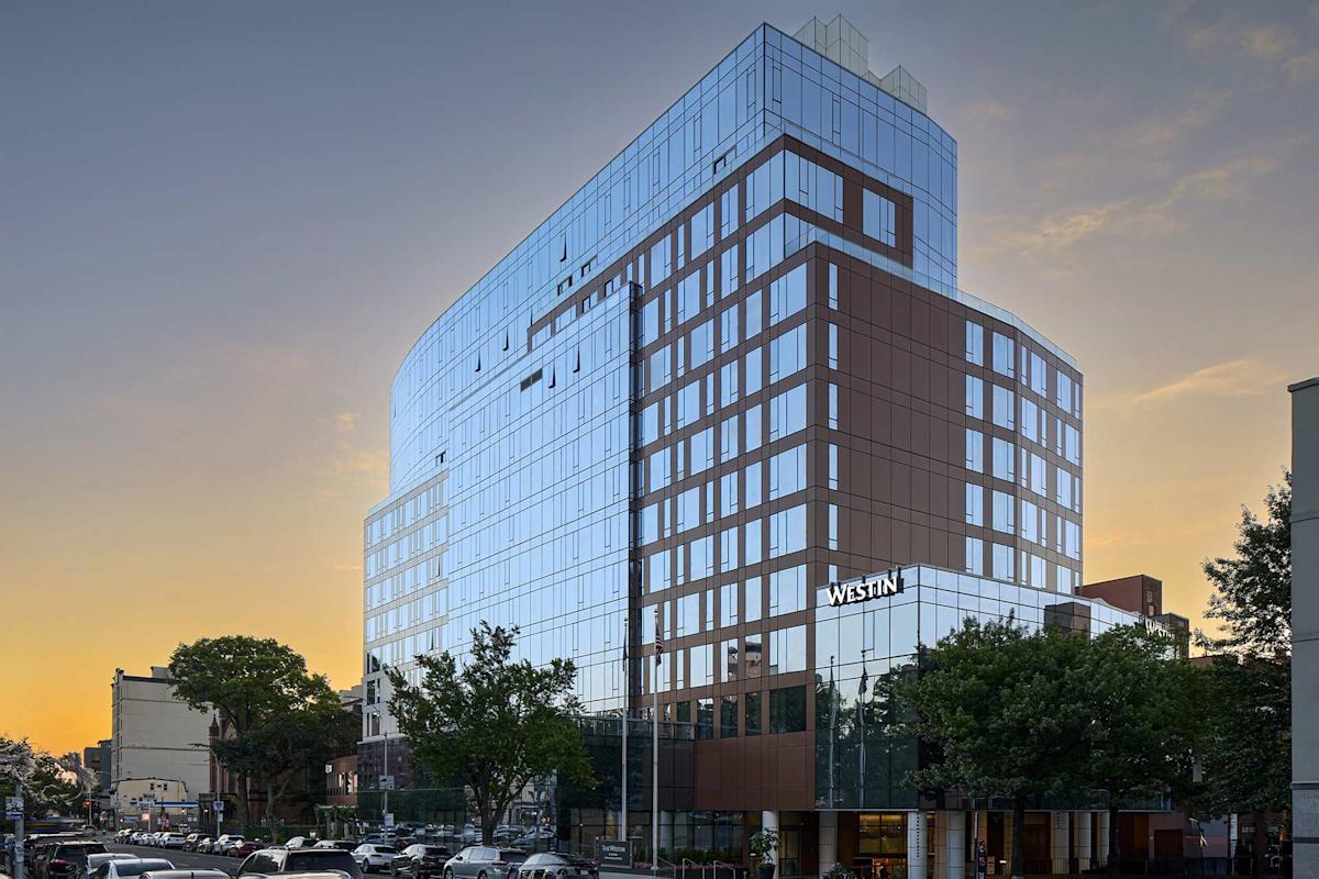 A modern, glass-fronted high-rise hotel building with "Westin" signage, set against a sunset sky. Trees and cars line the busy street in front of the hotel.