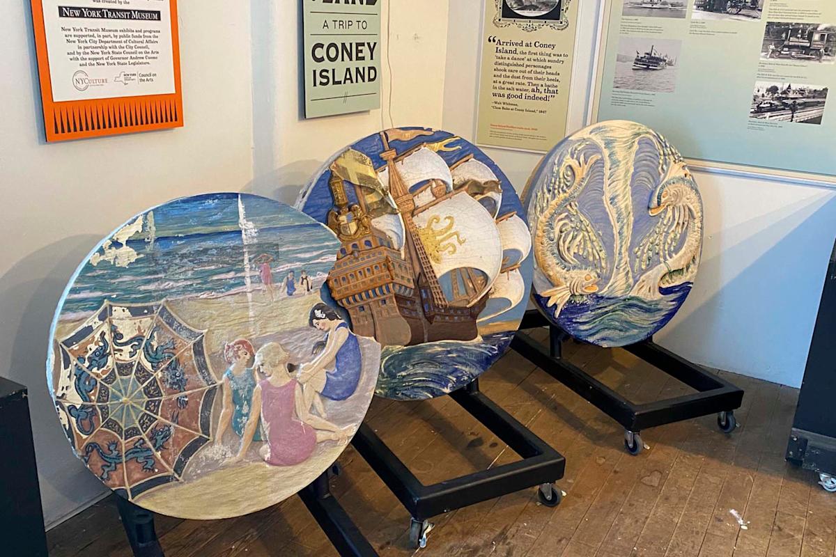 Three circular, intricately carved wooden panels are displayed on stands. They feature scenes of a beach gathering, a large ship with sails, and two sea creatures. Historical posters related to Coney Island are visible in the background.