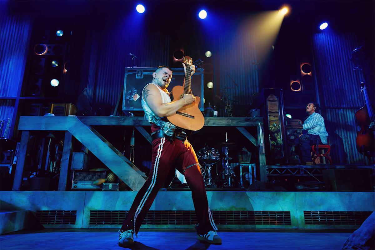A musician in red pants passionately plays a guitar on stage under blue and purple lights, while another person plays piano in the background amid an industrial-style set.
