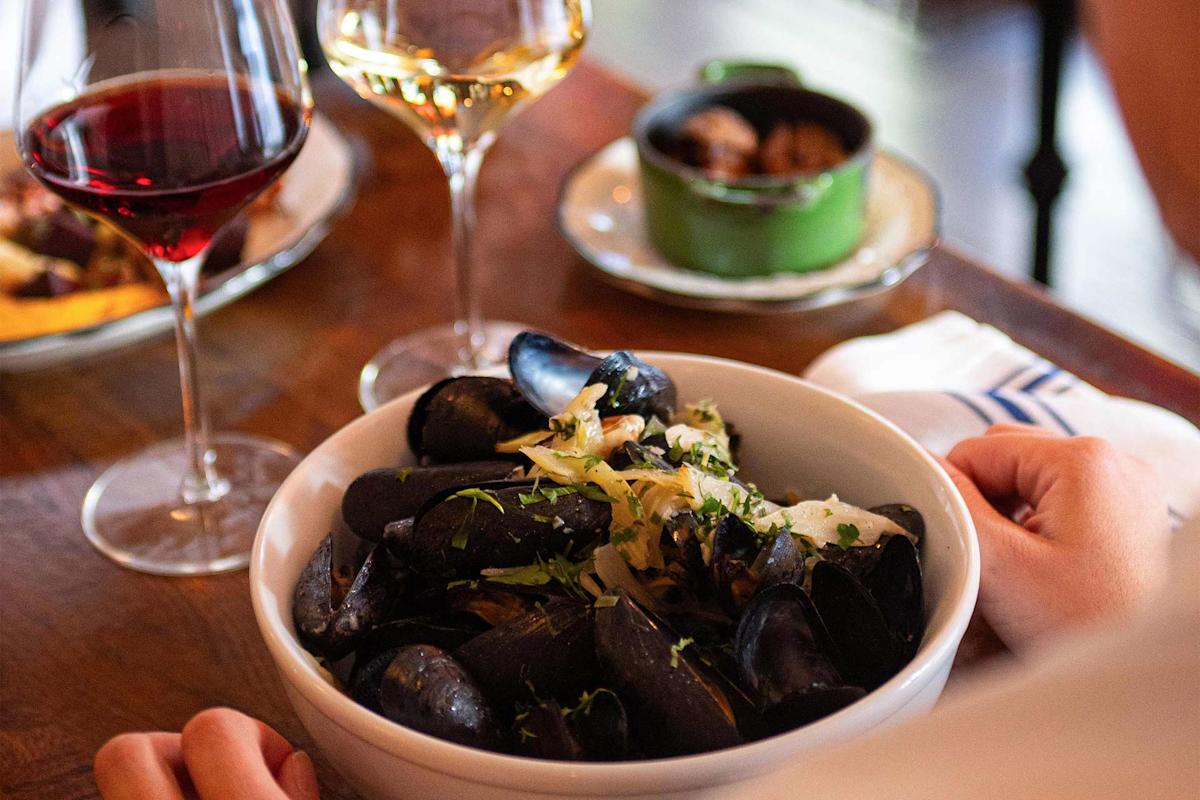 A person sits at a wooden table with a bowl of mussels garnished with herbs and onions, flanked by glasses of red and white wine and a small green pot of food in the background.