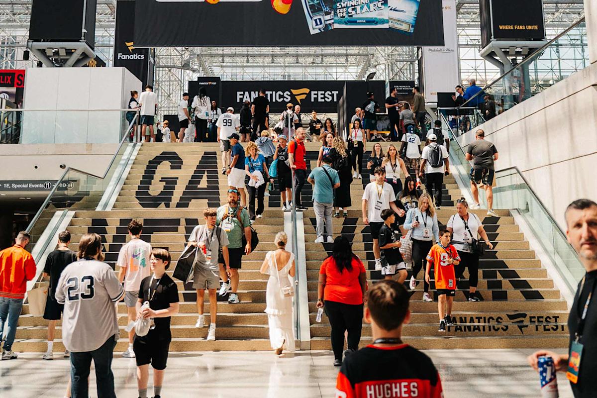 A busy indoor event with many people walking up and down wide stairs. Some wear sports jerseys and carry bags. Large signs above and on the stairs read “Fanatics Fest.” The area appears bright and spacious.