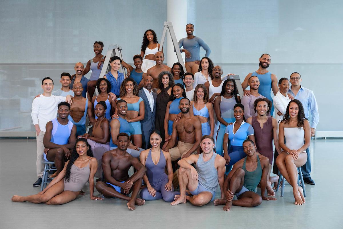 A large, diverse group of dancers pose together in a studio. They wear form-fitting dance attire in neutral and pastel tones, smiling and standing or seated in rows, with bright natural light in the background.