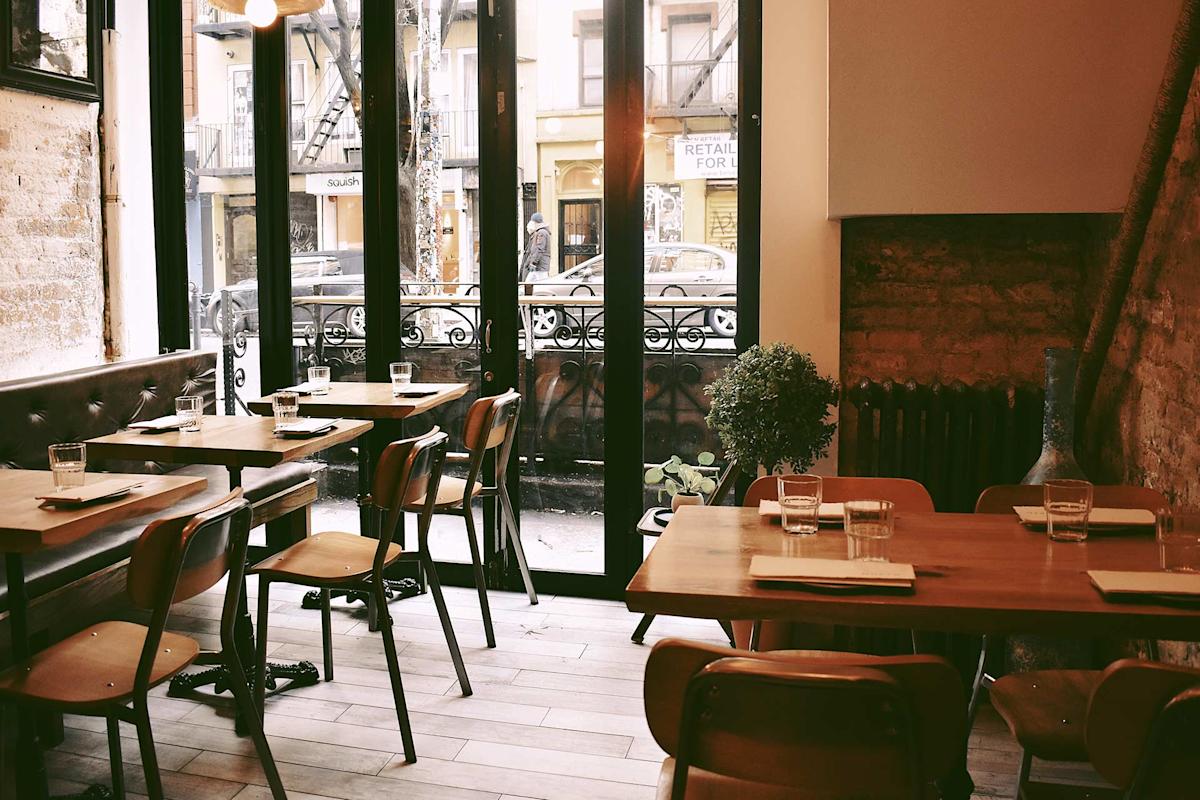 A cozy, sunlit café with wooden tables and chairs, set with glasses and napkins, next to large windows overlooking a city street and a small outdoor patio area.