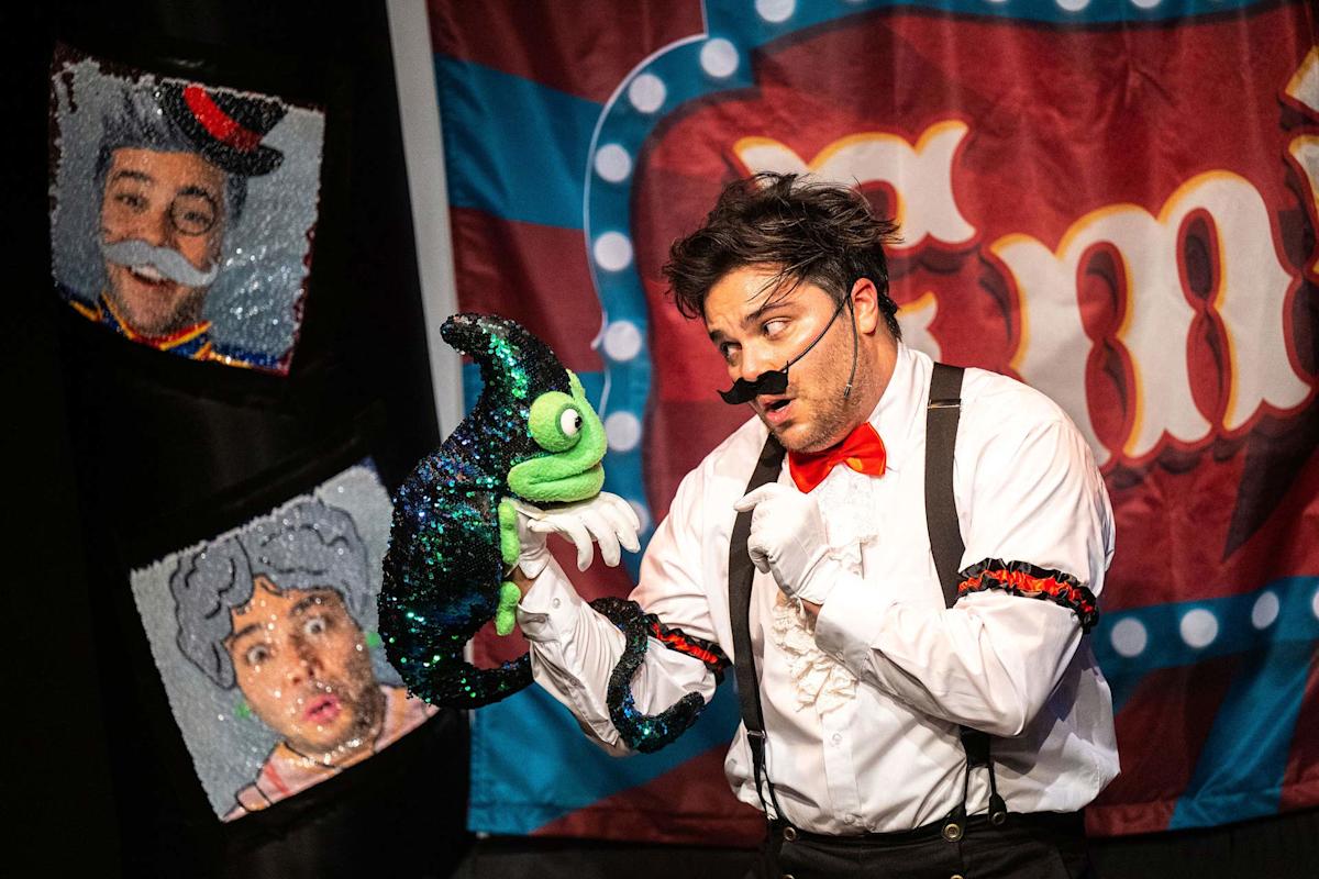 A performer dressed in a white shirt, red bow tie, and suspenders interacts with a colorful puppet. The background features a decorative banner with the letter "T" and sequined images of surprised faces.