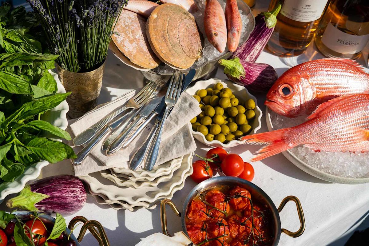 A table with fresh red fish, clams, eggplants, basil, green olives, tomatoes, cutlery, a dish of roasted tomatoes, bottles of wine, and a vase of lavender on a white tablecloth in bright sunlight.