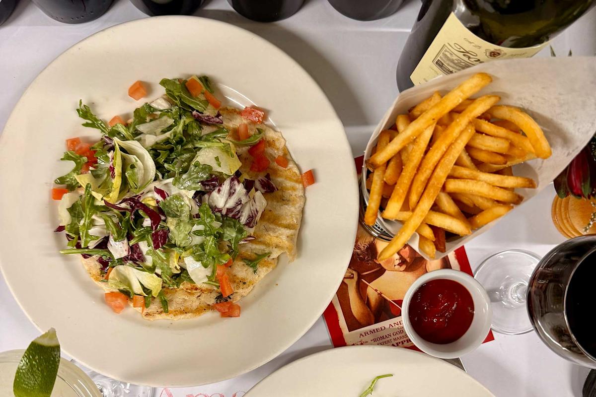 A plate with flatbread topped with mixed greens and diced tomatoes sits next to a container of crinkle-cut fries and a small dish of ketchup. Bottles of wine, a wine glass, and a lime wedge are also visible.