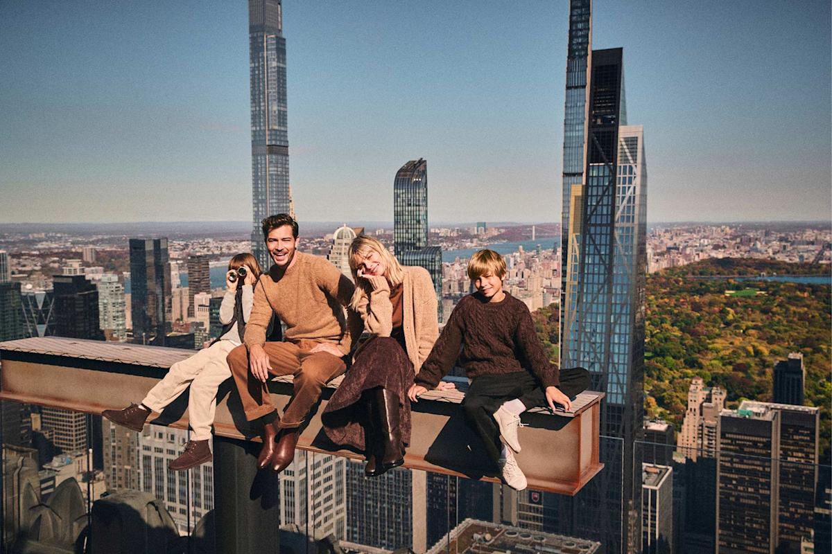A smiling family of four sits on a steel beam high above a city skyline with tall skyscrapers and a park below, posing for a photo in autumn clothing.