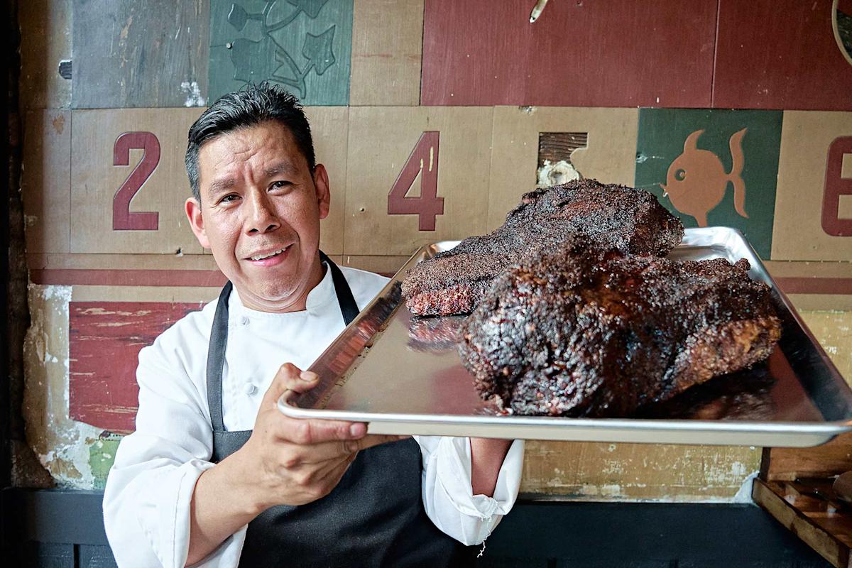 A chef in a white coat and black apron smiles, holding a tray with large pieces of smoked, seasoned meat in front of a colorful, rustic wall with numbers and designs.