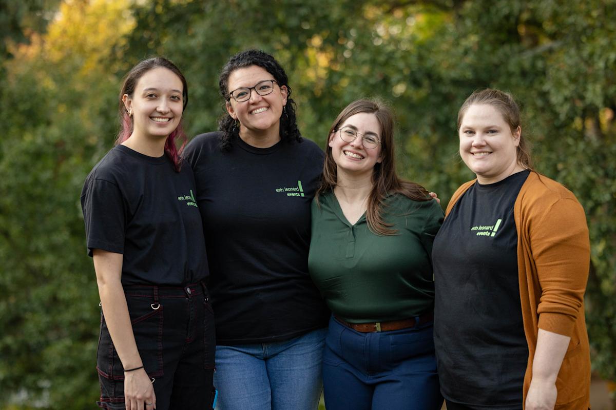 Four people stand outdoors smiling. The group includes three individuals in black shirts and one in a green blouse. The background is a lush, green forest.