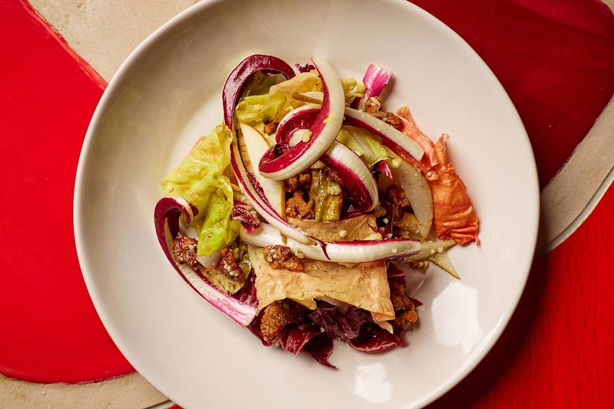 A white bowl filled with a mixed salad of leafy greens, radicchio, sliced onions, crispy croutons, and what appears to be walnuts, set on a table with red and beige placemats.