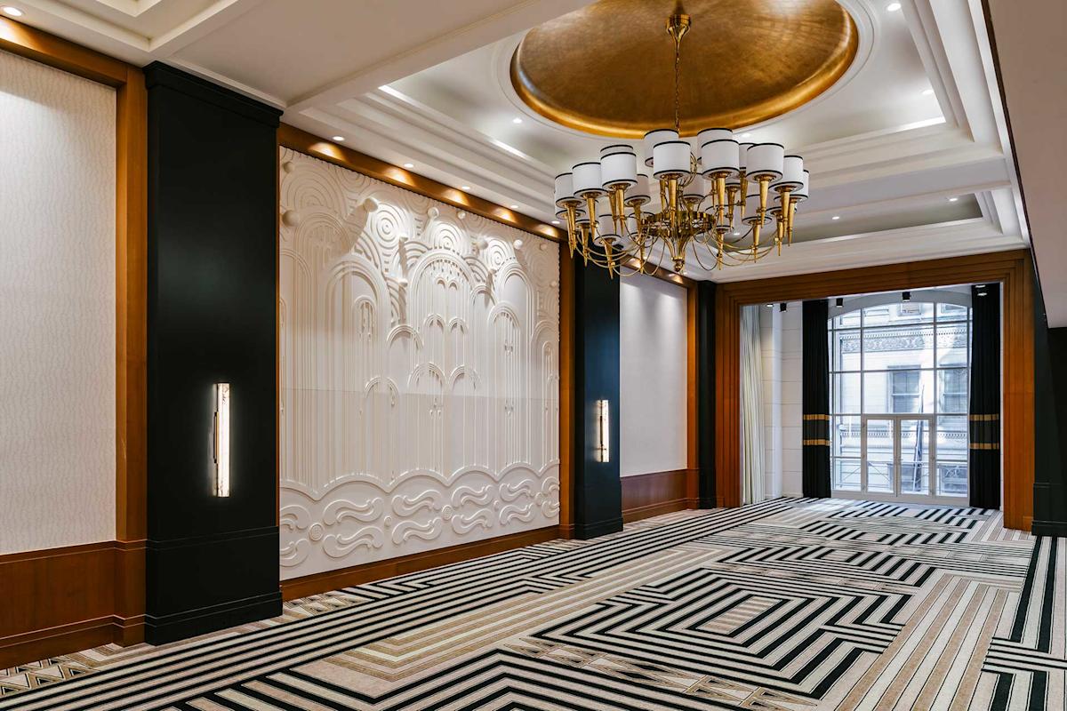 A modern, elegant hallway with a large gold chandelier, geometric black-and-white carpet, decorative white wall panels, wood accents, and large windows in the background.