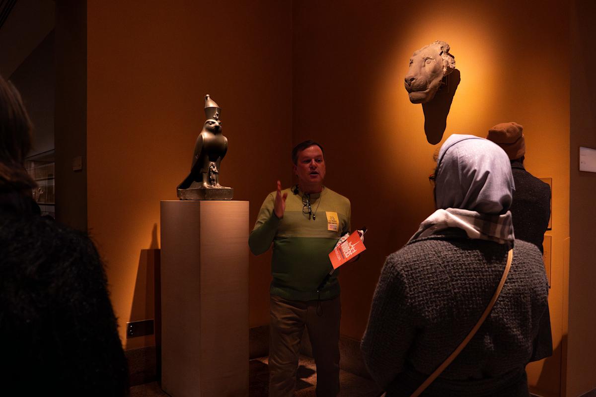 A museum guide gestures while speaking to visitors near ancient sculptures, including a falcon statue and a lion's head, displayed against a warmly lit orange wall.
