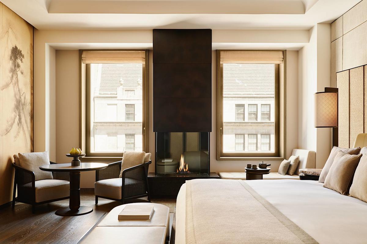 A modern, elegant hotel room with a large bed, sitting area, fireplace, tall windows with city views, and neutral-toned decor. Natural light fills the space, highlighting the cozy and minimalist design.