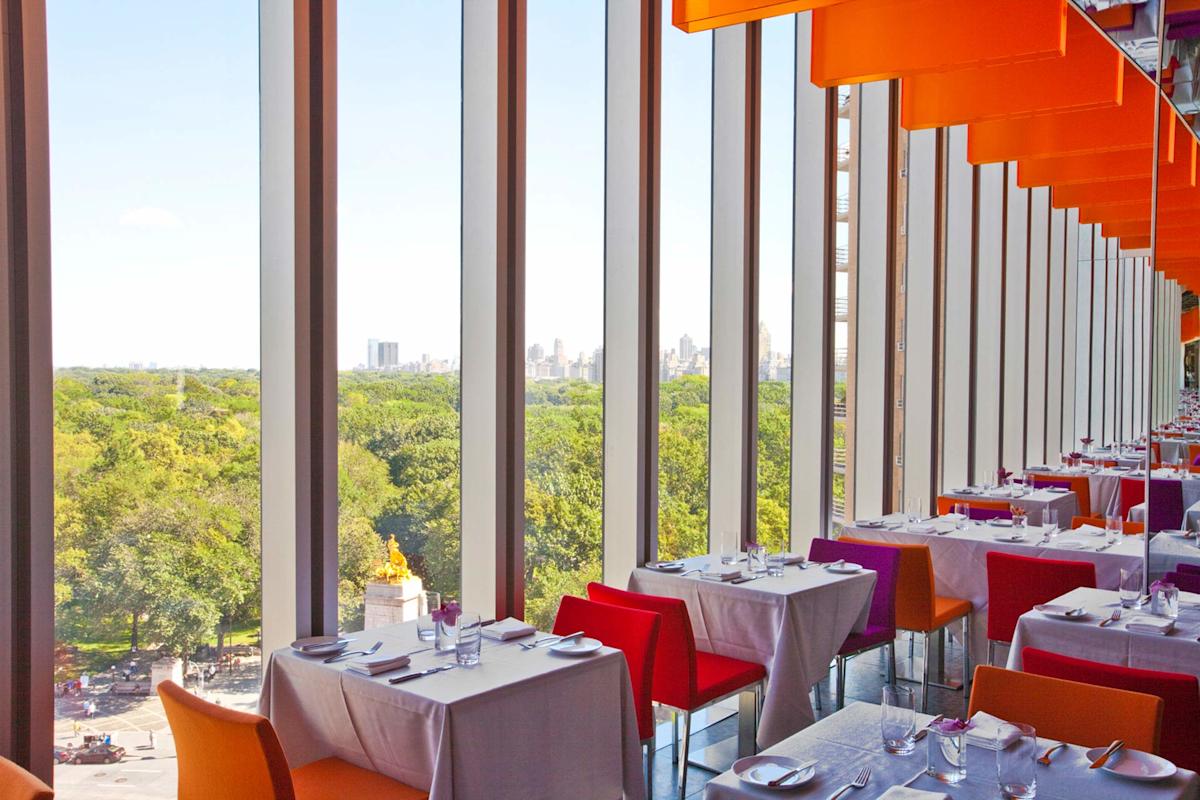 Modern restaurant with large floor-to-ceiling windows overlooking a green park and city skyline. Tables are set with white tablecloths and colorful chairs, and orange ceiling panels add a vibrant touch.