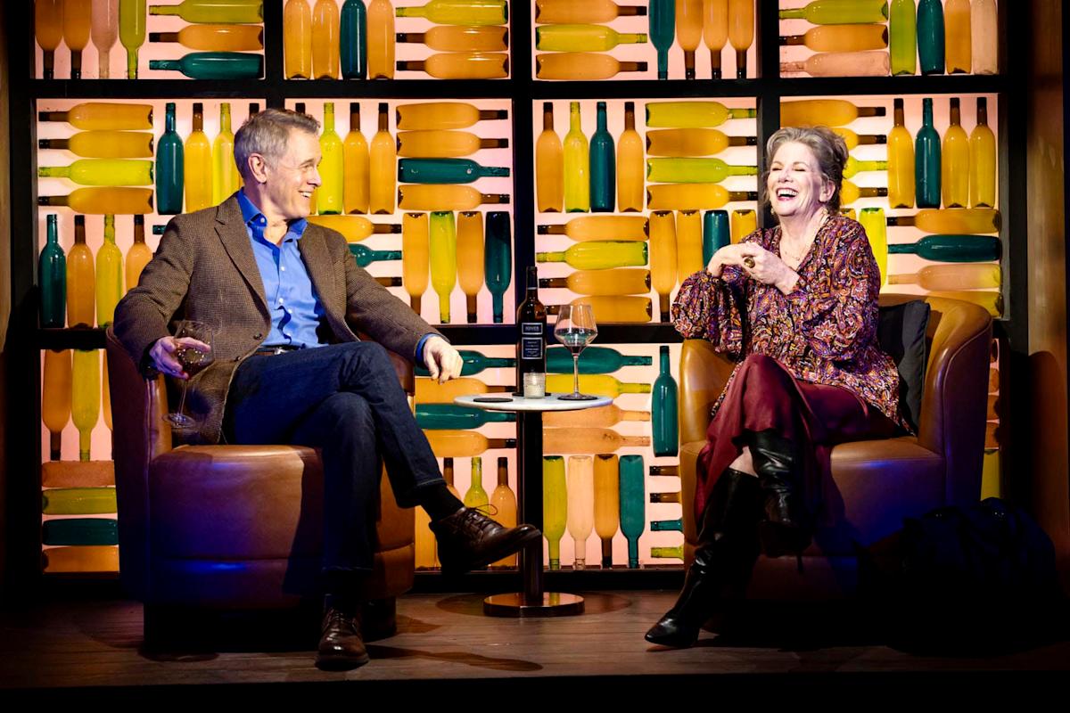 A man and woman are sitting in leather chairs on stage, laughing together. They each hold a wine glass. Behind them is a wall decorated with colorful bottles. A small table with a bottle is between them.