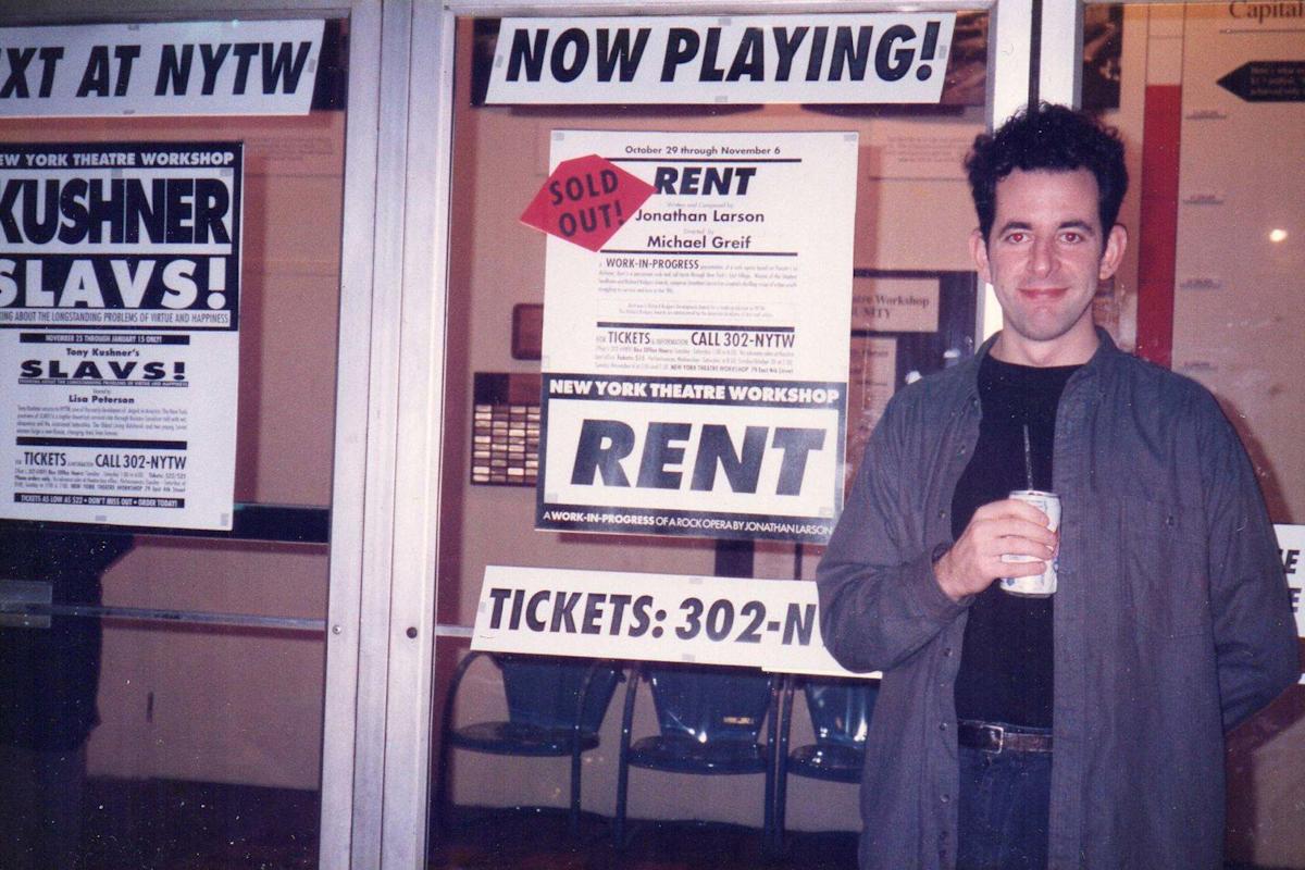 A person holding a drink stands in front of a theater displaying "Rent" posters, with "Now Playing!" and "Sold Out" signs. The background includes glass doors and additional theater advertisements.